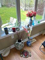 Photo of various glass and ceramic vases arranged on the floor and window sill including faux flowers and a Good Luck sign on the floor.