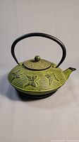 Green cast iron teapot with raised butterfly motifs, rounded lid, and black handle against neutral background.