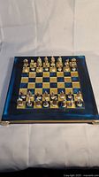 Full view of chess set with all pieces arranged on the board. Board is blue and gold metallic with 11x11 squares. Pieces are gold and silver metallic with Roman inspired design.