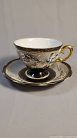 Front and side of teacup and saucer showing raised dragon design, black and gold detailing, and gold handle on teacup.