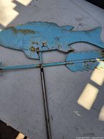 Blue fish-shaped metal weather vane with rust and paint wear, showing arrow and pointer mechanisms attached to a metal pole.