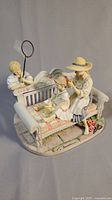Figurine showing child holding removable butterfly net and two girls on ornate bench with floral quilt cushion and grass base.