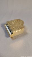 Closed view of the miniature brass piano keepsake box showing embossed floral design and keyboard detail.