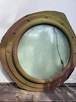 Front view of weathered metal porthole with cracked greenish glass, showing algae and rust on surface.