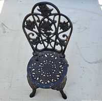 Front view of small ornate cast iron doll-sized chair showing decorative leaf motif backrest and circular floral seat with three curved legs.