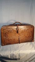 Full front view of vintage tooled leather suitcase showing worn leather and central buckle strap.