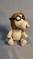 Front view of Snoopy figure seated, showing white body, brown aviator hat with black goggles, and paint wear on nose and limbs.