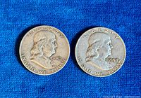 Two American half dollar coins on blue fabric, displaying obverse side with Benjamin Franklin's profile and dates 1952 and 1958.