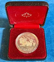 Coin in protective capsule displayed inside red velvet clamshell presentation case with maple leaf insignia on lid interior.