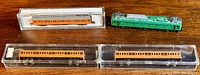Three orange passenger train cars in clear plastic cases and one green locomotive engine on wooden surface.