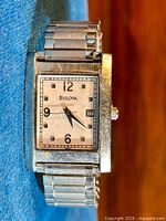 Front closeup image of watch showing rectangular face, dial with Arabic numerals 12, 3, 6, 9 and date window, stainless steel band.