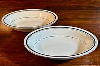 Two identical white Minton's 'Bridal Veil' fine bone china oval serving bowls on a wooden surface, showing the overall design and shape of the bowls with black trim and embossed interior pattern.