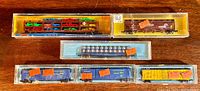 Overview photo showing six N gauge train cars including a multilevel autorack with cars, brown boxcar, blue hopper, yellow specialty boxcar, and two blue Atlas boxcars in original packaging.