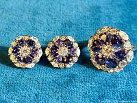Photo showing two floral stud earrings and one matching floral ring, all made of sterling silver with blue and clear cubic zirconia stones arranged in a flower pattern, placed on blue fabric.