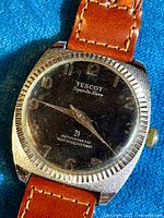 Close-up photo showing black dial with gold-tone numbering, brushed metal case with fluted edge, and stamped inscriptions on dial.