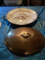 Porcelain serving tray with floral and bird decoration, dark handles on either side, shown with copper warming plate lid on top