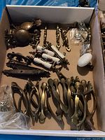 Box containing multiple sets of antique dresser hardware including brass and porcelain handles and pulls arranged in an open cardboard box.