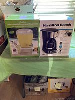 Both product boxes side by side on a green tablecloth, Hamilton Beach coffee maker and 3 gallon beverage dispenser.