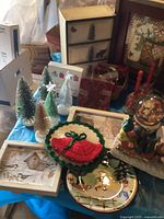 Photo shows various Christmas decorations including bottle brush trees, crocheted Christmas doilies, decorative plates and trays, and a wooden tiered box in the background.