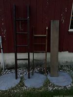 Two wooden decorative ladders and a long piece of barn wood placed on gray concrete pads against red barn wall.