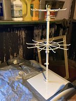 White metal swivel jewelry display stand with multiple horizontal arms for hanging jewelry or small items, placed on a white surface with paint cans and tools in background.