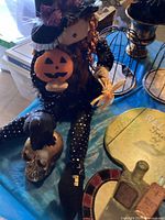 Image showing witch doll sitting on table holding pumpkin decoration, skull with black hat nearby, spider decoration visible as well.