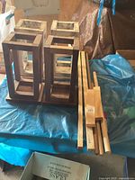 Four handmade wooden lantern frames, two with natural wood tone and two whitewashed, shown on a tarp-covered surface.