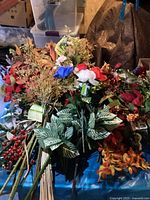 Large assortment of artificial flowers on a surface, including red, white, blue flowers, variegated green leaves, and orange seasonal stems with berries.