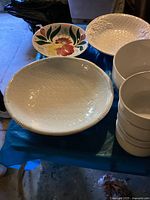 Wide view showing large basket weave patterned ironstone plate, colorful floral plate, large white salad bowl, and four small white bowls stacked