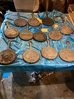 Photo showing 12 heavy metal Pennsylvania Dutch style trivets arranged on a blue cloth, displaying embossed historical buildings and dated motifs.
