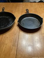 Two Ruff Hewn cast iron skillets on a wooden table; left skillet is larger (10 inch), right skillet is smaller (7 inch). Both have solid handles and classic black finish.