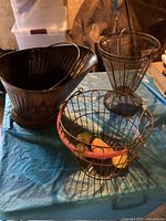 Three vintage baskets grouped on a table: a black tin bucket with handle, a tall wire plant holder, and a wire egg basket with wooden rim and faux fruit inside.