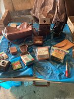 Full view of vintage camera lot with cameras, leather bags and boxes of film and flash bulbs on blue tarp table.