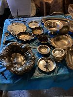 Overview of various vintage silver and silver-plated dishes and bowls on a blue cloth, showing different types of serving pieces including a double-tier stand and a large decorative bowl.