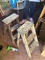 Two small wooden ladders side by side on floor, showing peeling white paint and worn condition, legs, steps, and surrounding items visible.