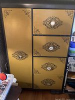 Front view of yellow wood cabinet with four drawers and one side armoire door, showing black decorative patterns and glass knobs.