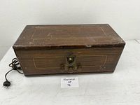 Wooden box-shaped radio from front side with power cord, shows scratches on wooden exterior and brass metal hardware.