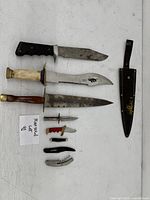 Multiple cutting utensils laid out against a white background including various fixed and folding knives, plus a sheath and corkscrew.