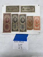 Front view of seven foreign paper money notes mounted on a clear acrylic display frame secured with metal screws at corners.