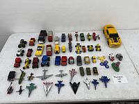 Top-down view showing the full collection of small toys cars, trucks, planes, two fidget spinners and a green toy figure arranged on a white surface.
