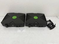 Front and side view of two original Xbox consoles placed side by side on a white surface with a handwritten sign 'MaxSold Lot 37'.