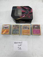 Four stacks of pokémon trading cards with gray borders in sleeves, each set topped with a different card, displayed in front of a black metal Pokémon tin storage box featuring Yveltal design.
