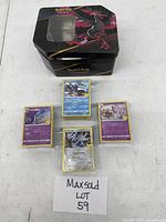Black tin storage box decorated with Pokémon artwork, shown with four stacks of sleeved Pokémon cards placed in front, labeled 'Maxsold Lot 59'.