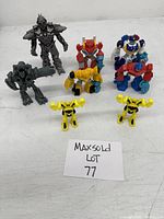 Lot of seven miniature Transformer toy figures arranged on white surface including two yellow Bumblebee, red and blue Optimus Prime, blue and white police robot, and two grey Decepticon resembling Megatron.