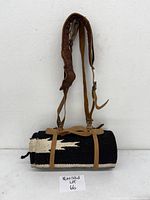 Rolled black and white Navajo saddle blanket strapped with woodrich leather carrying strap hung against white wall