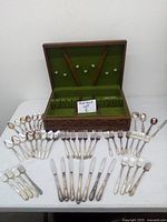 Opened wooden case with green felt interior holding arrangement hooks and compartments. Tarnished vintage Rogers silverware displayed in front including forks, knives, spoons, and butter knives.