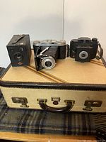 Lot of 3 antique cameras arranged on a vintage suitcase with a plaid fabric underneath, showing all three cameras together.