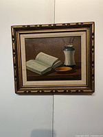 Full view of the framed still life painting depicting an open book, a vase, and a saucer.