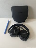 Black Bose wired headphones with cushioned ear pads that show wear, paired with a black zippered carrying case with Bose logo, placed on a white surface next to a blue pen for scale.