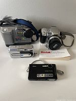 Four digital cameras displayed: Sony, Kodak, Nikon Coolpix, and Fujifilm. Kodak manual visible.